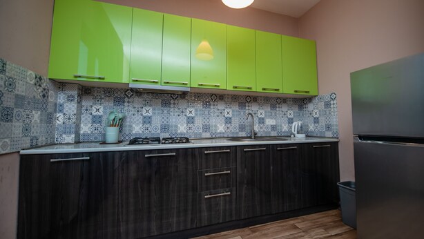 Een keuken met groene kastjes, een betegelde spatwand, een roestvrijstalen koelkast en een houten vloer.