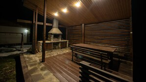 Een terras buiten met een stenen vloer, houten banken, een stenen open haard en een houten plafond.