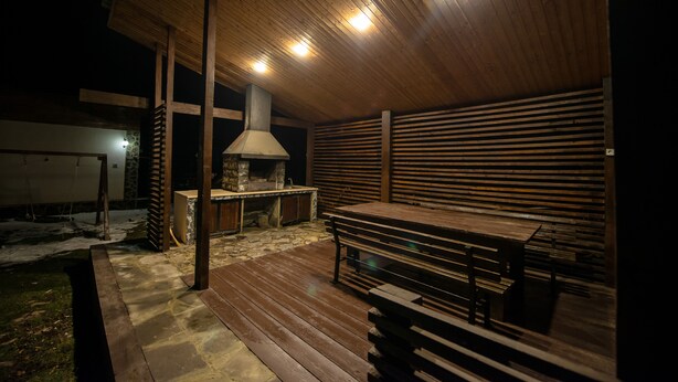 An outdoor patio with a stone floor, wooden benches, a stone fireplace, and a wooden ceiling.