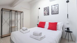 Ein ordentlich bezogenes Bett mit weißen und roten Kissen, ein Paar gefaltete Handtücher und ein kleiner, schwarzer Tisch mit einer Lampe.