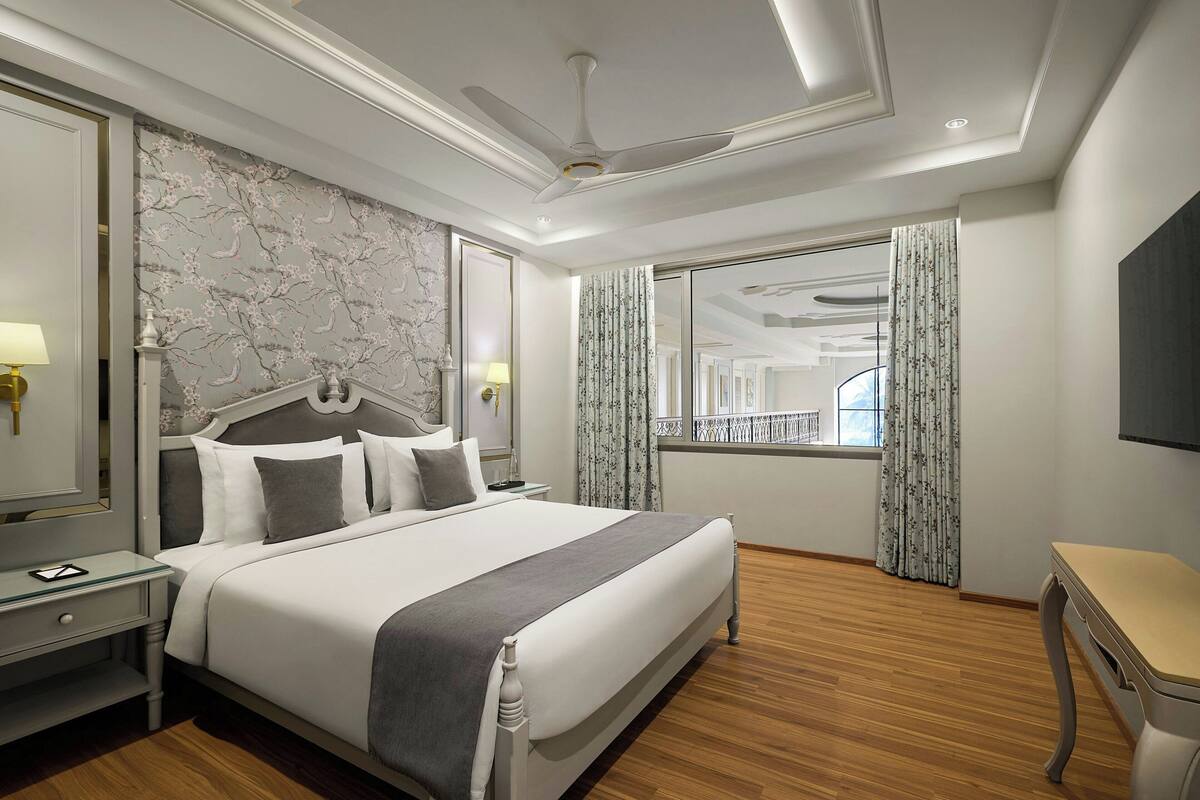 Ein Hotelzimmer mit einem großen Bett, einem Nachttisch, einem Schreibtisch, einem Deckenventilator und einem Fenster mit Vorhängen.