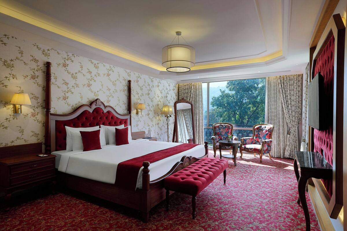 Ein Hotelzimmer mit einem großen Bett, einer Bank, einem Sessel und einem Tisch. Ein Fenster mit Vorhängen bietet Blick auf Bäume.