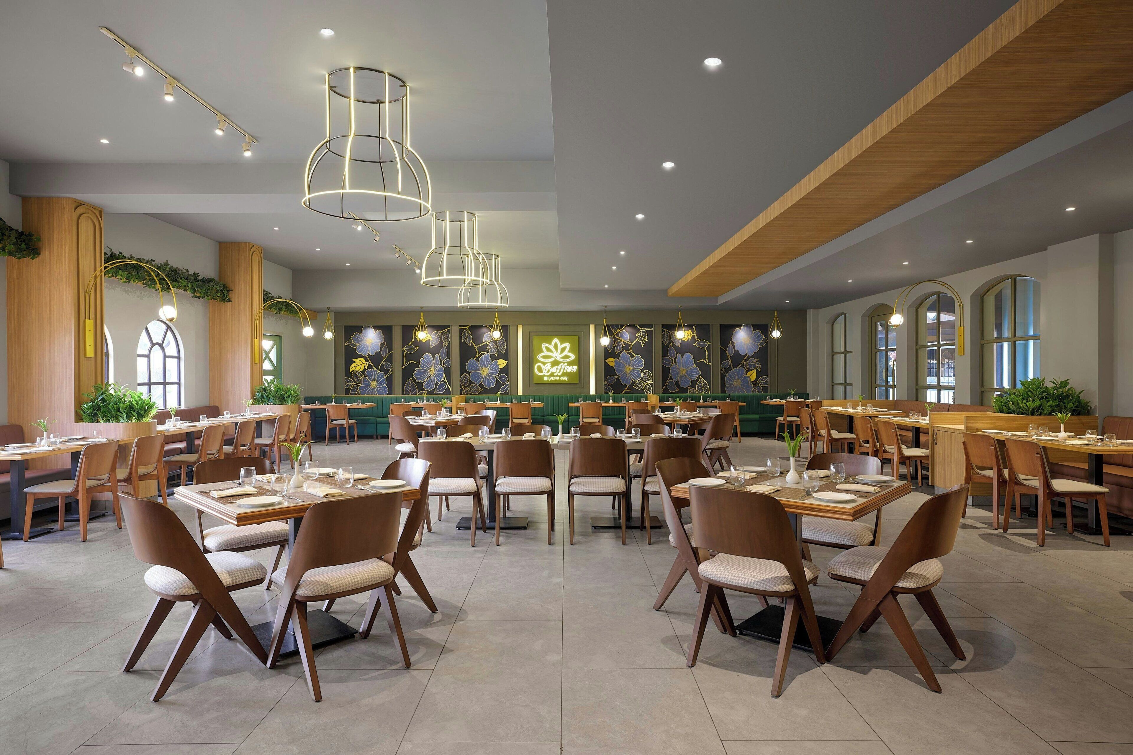 A spacious restaurant with multiple dining tables and chairs, modern pendant lighting, and a green wall with floral decorations.