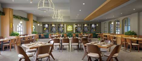 A spacious restaurant with multiple dining tables and chairs, modern pendant lighting, and a green wall with floral decorations.