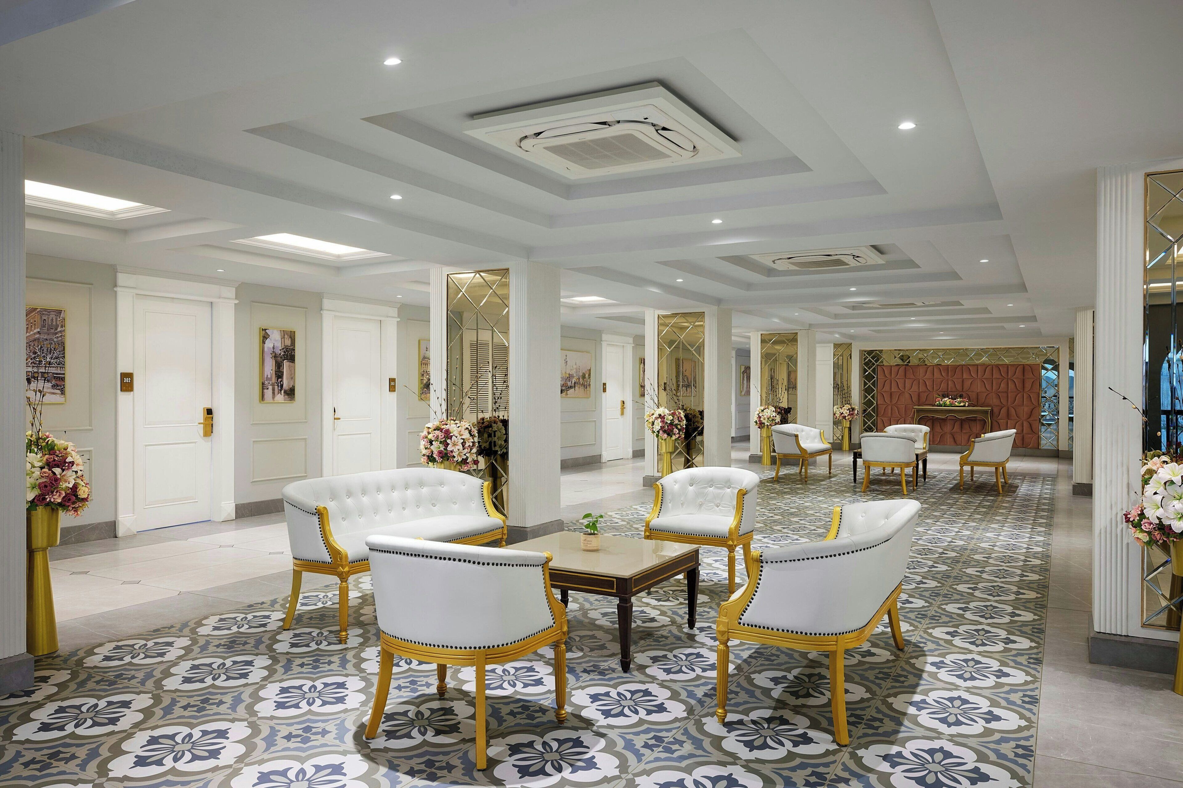A spacious hotel lobby with a patterned floor, white and gold upholstered chairs, and a central table.
