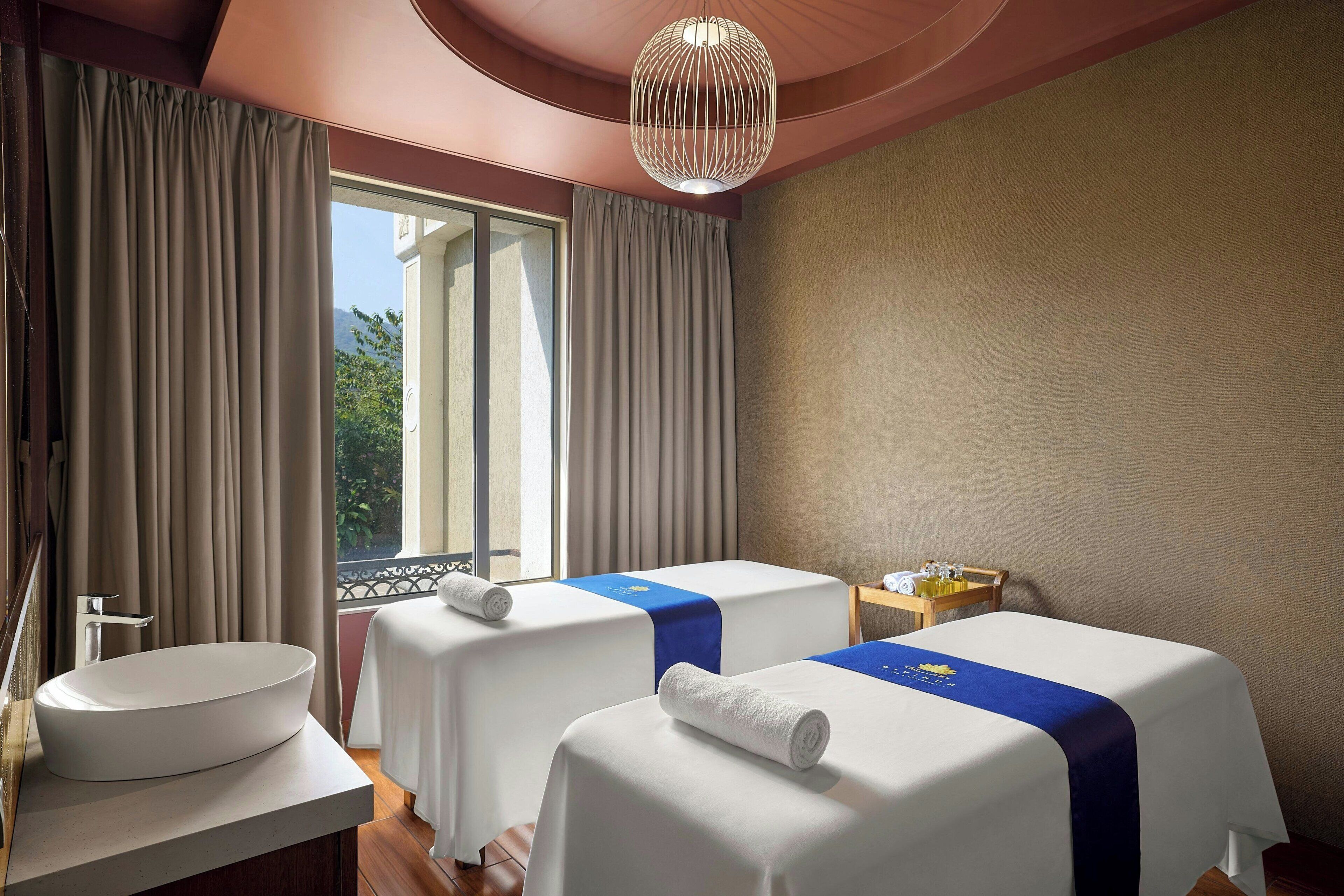 A hotel room with two beds, a large window with curtains, a sink, and a small table with toiletries.
