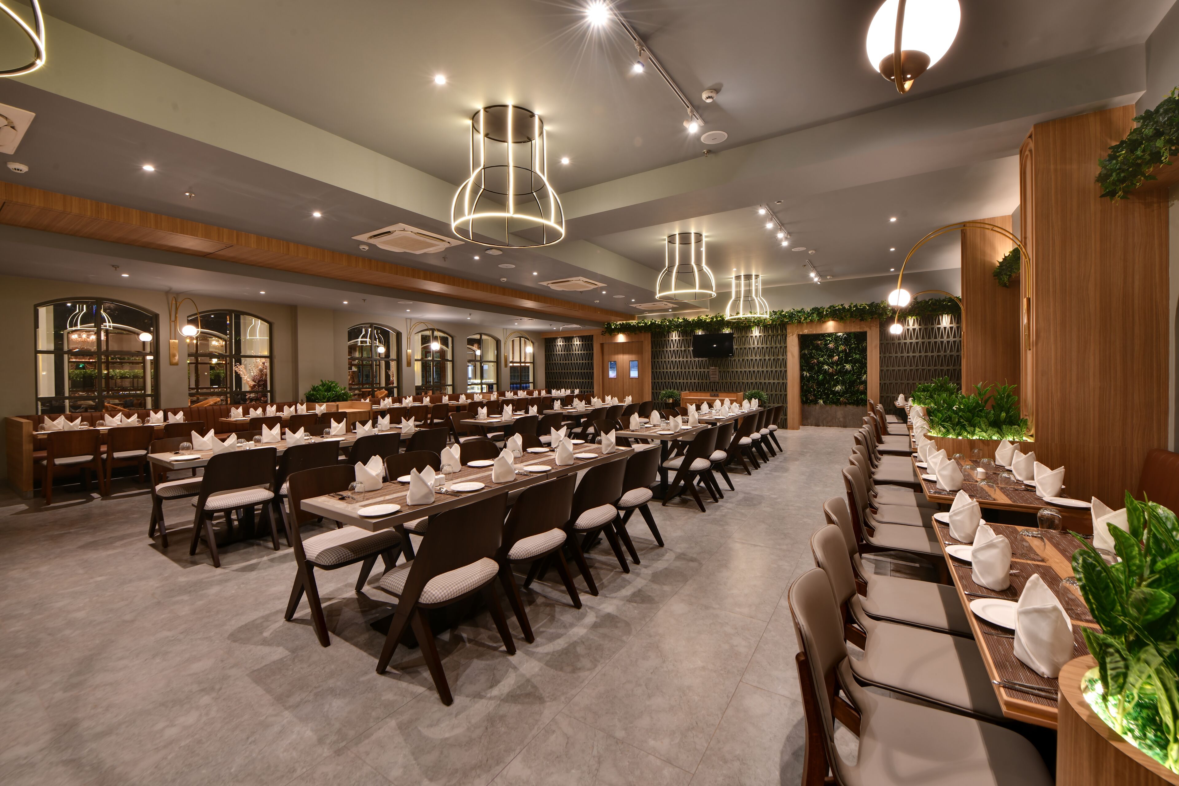 Une grande salle de banquet avec de longues tables dressées pour le repas, un éclairage moderne suspendu et un mur décoratif en bois agrémenté de plantes.