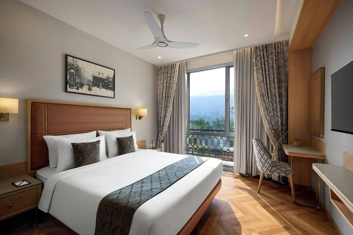 Ein Hotelzimmer mit einem großen Bett, einem Schreibtisch, einem Sessel, einem Deckenventilator, einem Fernseher und einem Balkon mit Vorhängen.