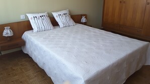 Ein ordentlich bezogenes Bett mit weißer Steppdecke, zwei Kissen mit Streifenmuster, ein Nachttisch aus Holz mit Lampe und ein Holzschrank.