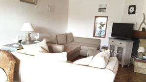 Ein Wohnzimmer mit einem beigefarbenen Ecksofa, einem Fernseher, einer kleinen Kommode und einem Korbstuhl.