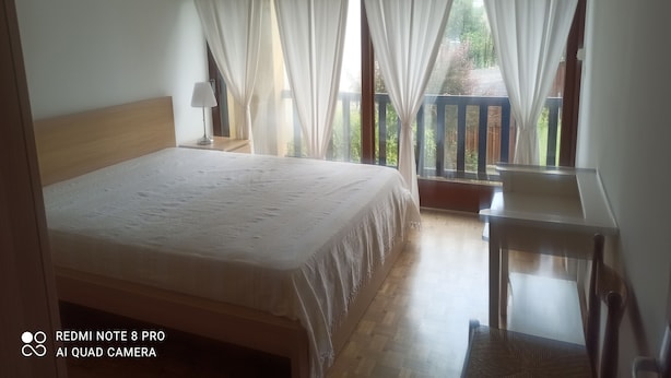 Ein Schlafzimmer mit Bett, Nachttisch, Lampe, einem Fenster mit Vorhängen und einem Balkon.