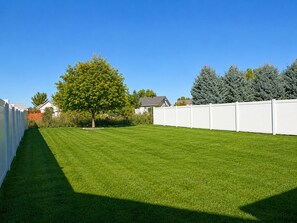 A well-maintained lawn with a white fence, a tree, and a row of evergreen trees in the background.