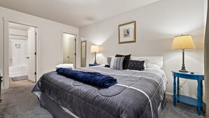 Et hotelværelse med en pænt redt seng, et natbord med en lampe, et billede på væggen og en dør til et badeværelse.