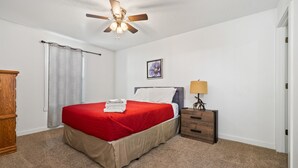 Un dormitorio con cama, mesita de noche, ventilador de techo, ventana con cortinas y cómoda de madera.