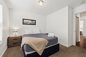 Ein Schlafzimmer mit einem Bett, einem Nachttisch, einer Lampe, einem gerahmten Bild an der Wand und einer Tür zu einem anderen Raum.