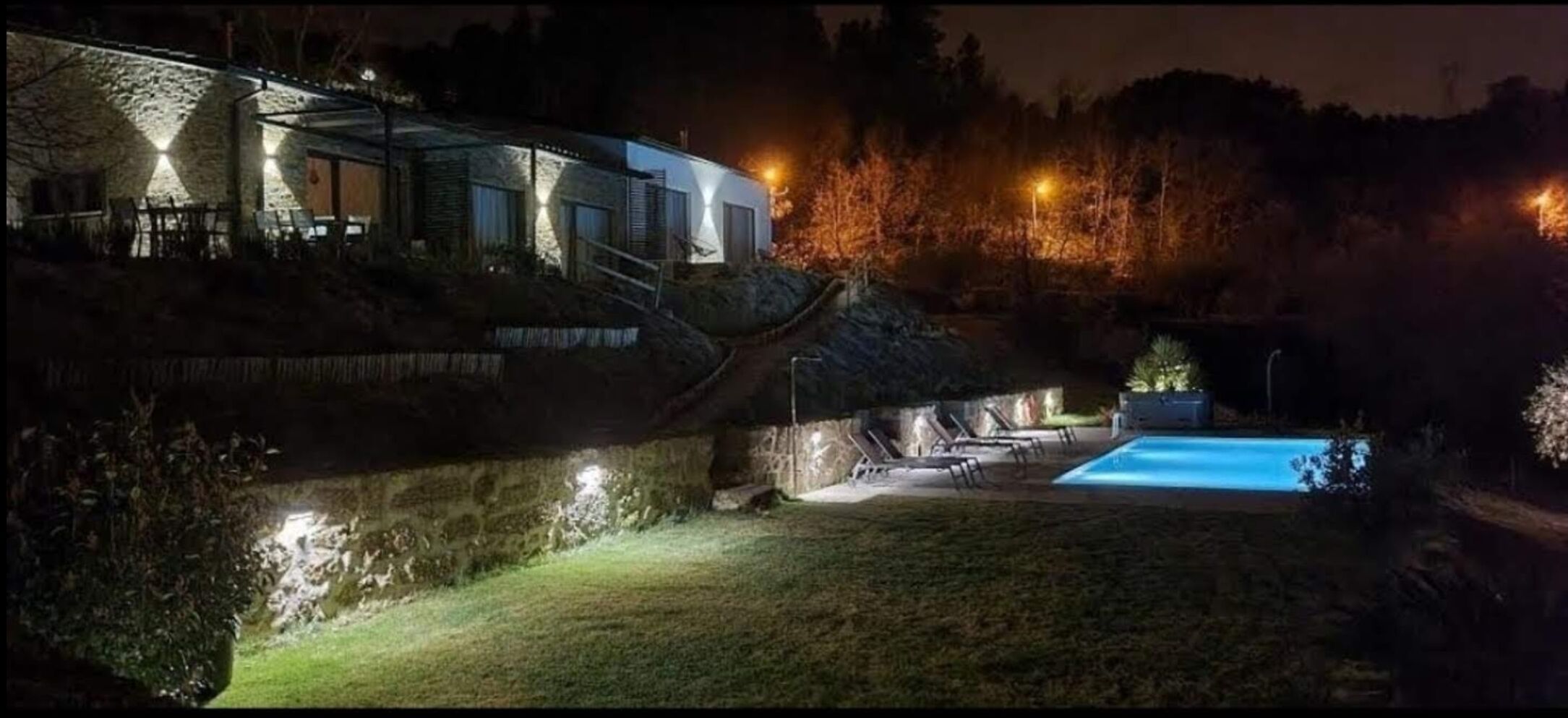 A nighttime view of a house with a swimming pool, stone walls, and outdoor lighting.