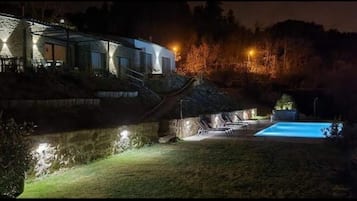 A nighttime view of a house with a swimming pool, stone walls, and outdoor lighting.