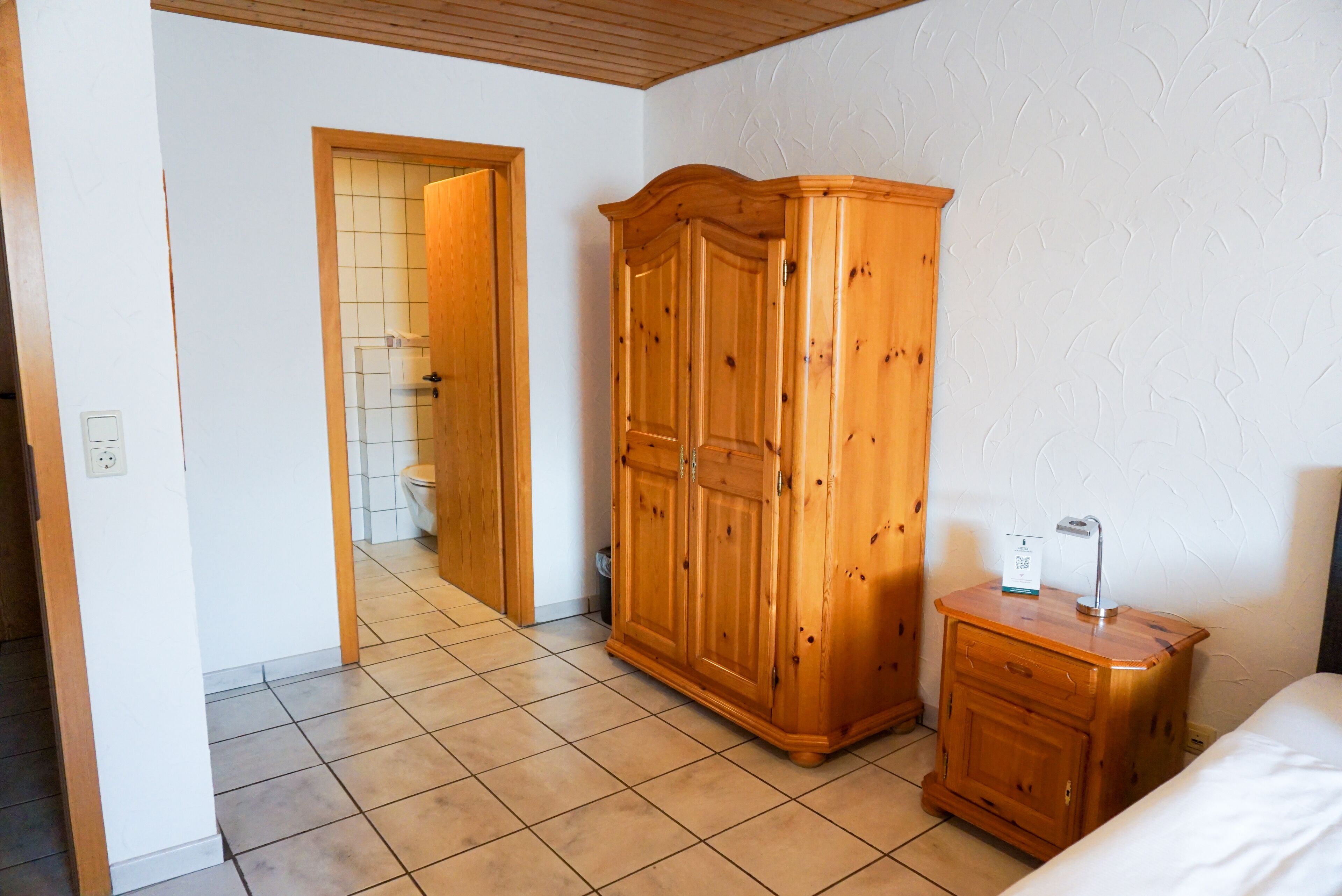 A bedroom with a wooden wardrobe, a bedside table, and a bed.