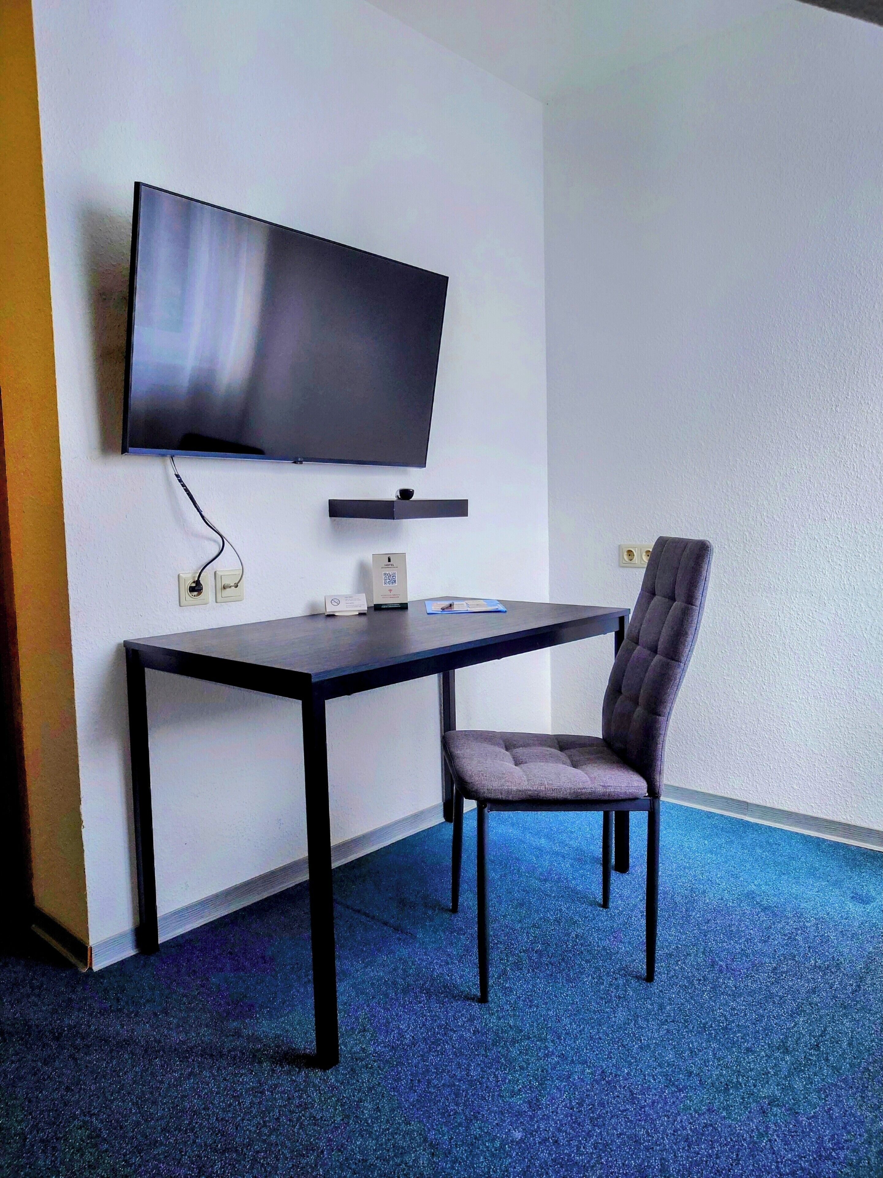 Ein Zimmer mit einem an der Wand befestigten Flachbildfernseher, einem dunklen Holztisch, einem gepolsterten Sessel und einem Teppichboden in Blau.
