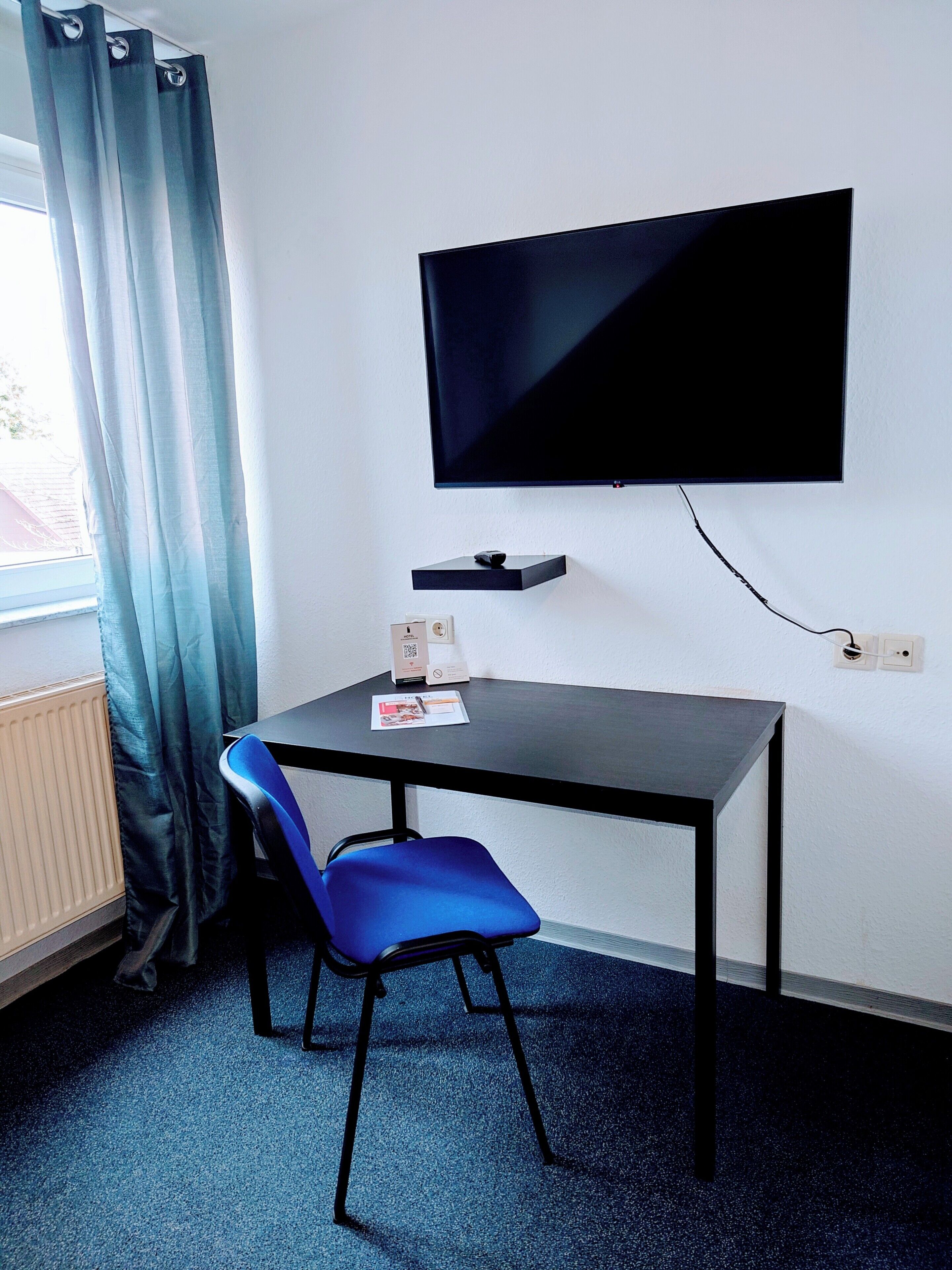 Una habitación con una televisión montada en la pared, una mesa con una silla, un radiador y una ventana con cortinas.