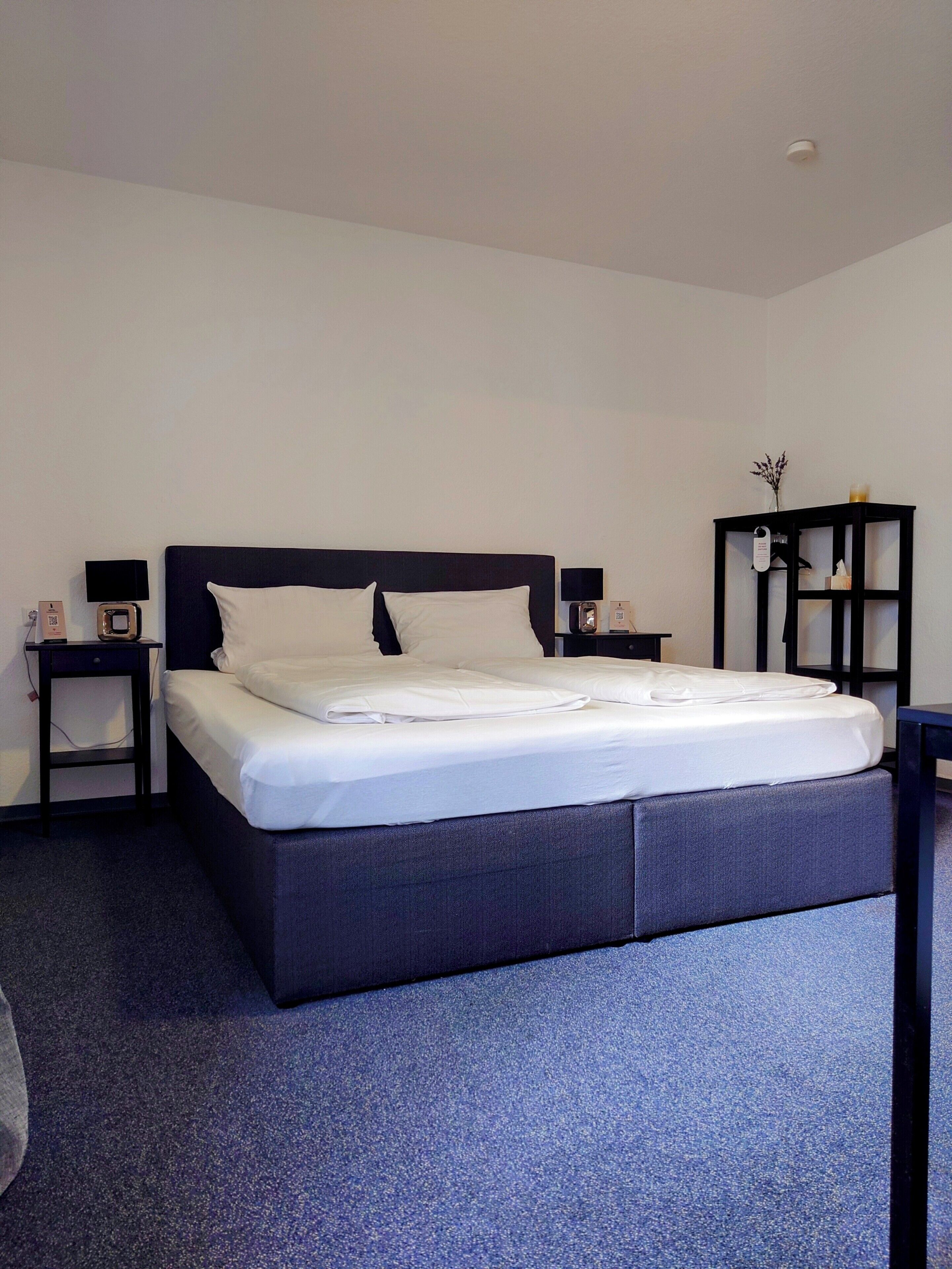 A neatly made bed with white linens, a dark headboard, and a small bedside table with a lamp and a clock.