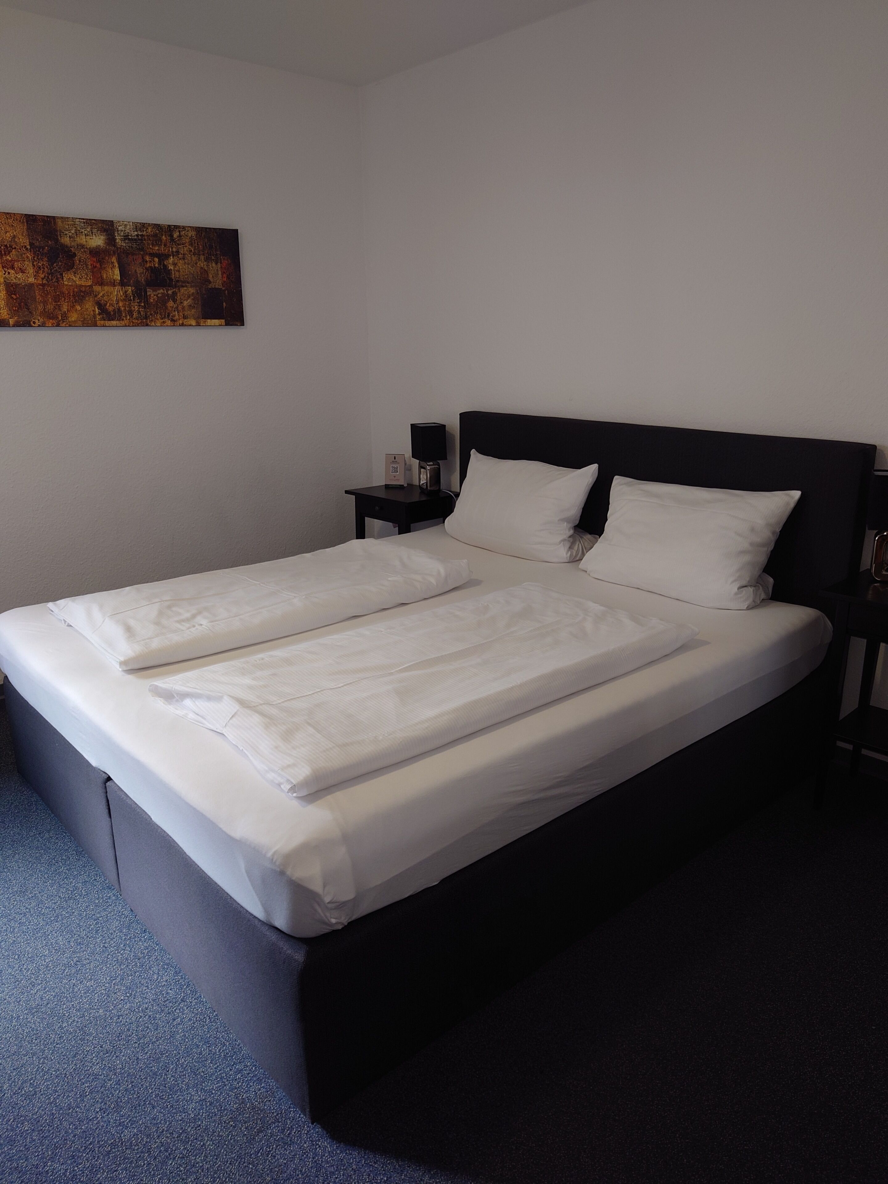 A neatly made bed with white linens, a black headboard, and a nightstand with a lamp and a small item on it.
