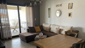 A modern living room with a sofa, a wooden dining table, and a balcony with a chair. - Beautiful apartment in the center of Folelli (Penta-di-Casinca)