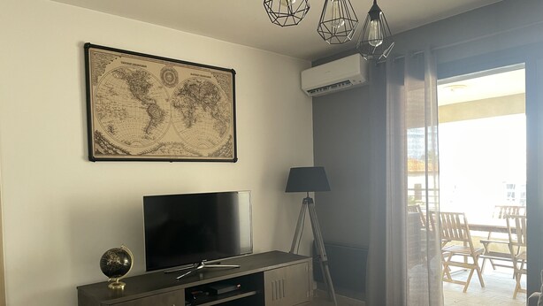 A living room with a flat-screen TV, a world map, a lamp, and a balcony with outdoor furniture. - Beautiful apartment in the center of Folelli (Penta-di-Casinca)