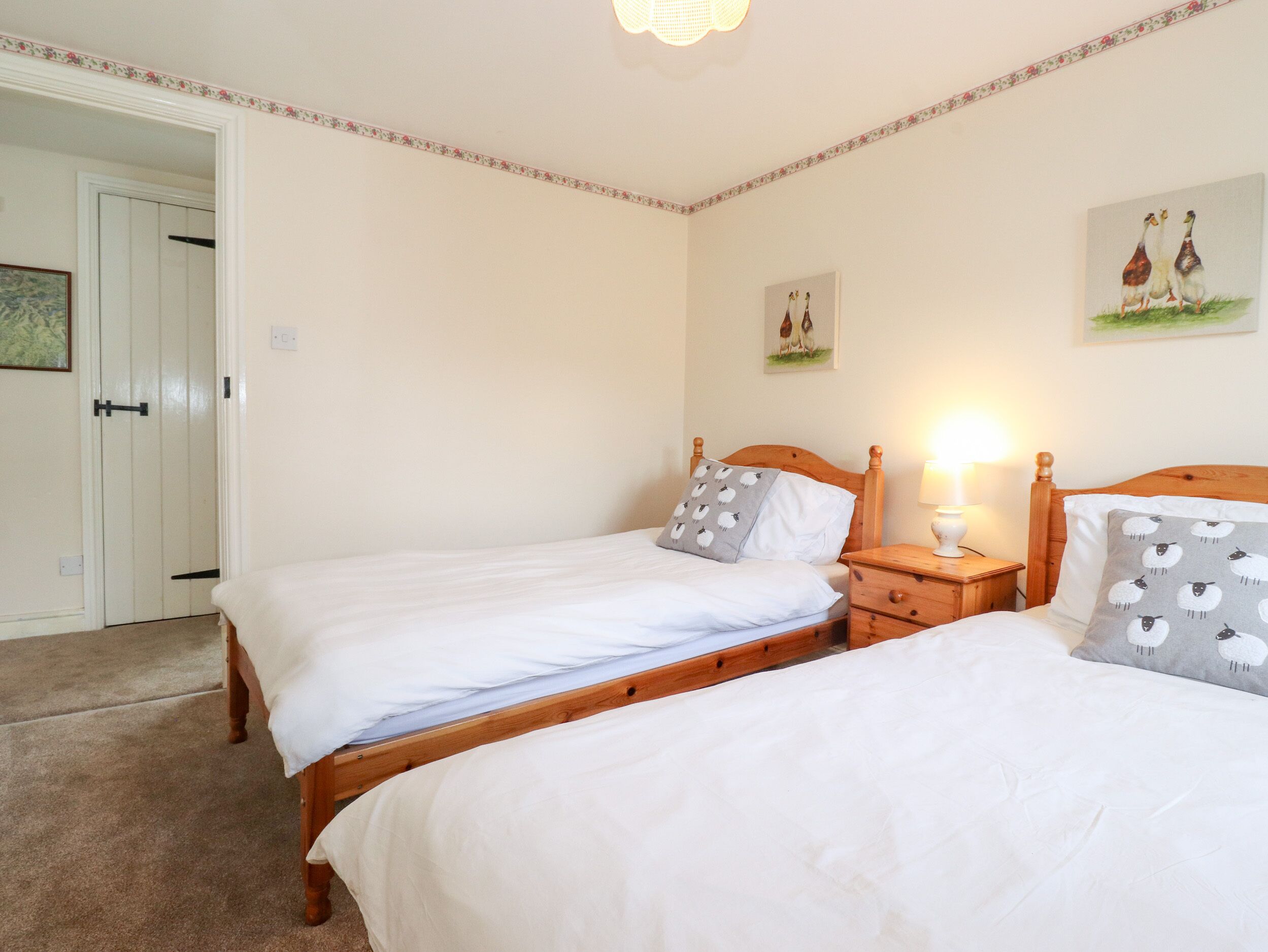 A bedroom with two single beds, wooden headboards, a bedside table with a lamp, and a door leading to another room.