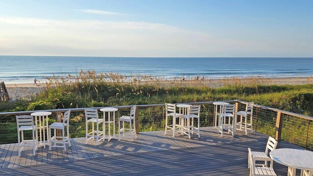 Terraza de madera con sillas y mesas blancas con vista a la playa.