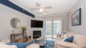 A living room with a fireplace, a ceiling fan, a large window with a view, and a sofa with cushions. - Casa Del Mare, beautiful 3 BD /3 Bathroom condo just steps to the beach, walking distance to activities. (Ocean City)