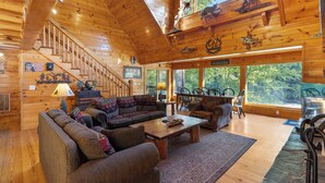 Interior de una cabaña de madera con una escalera, un sofá grande, una mesa de comedor y vistas al bosque a través de amplias ventanas.