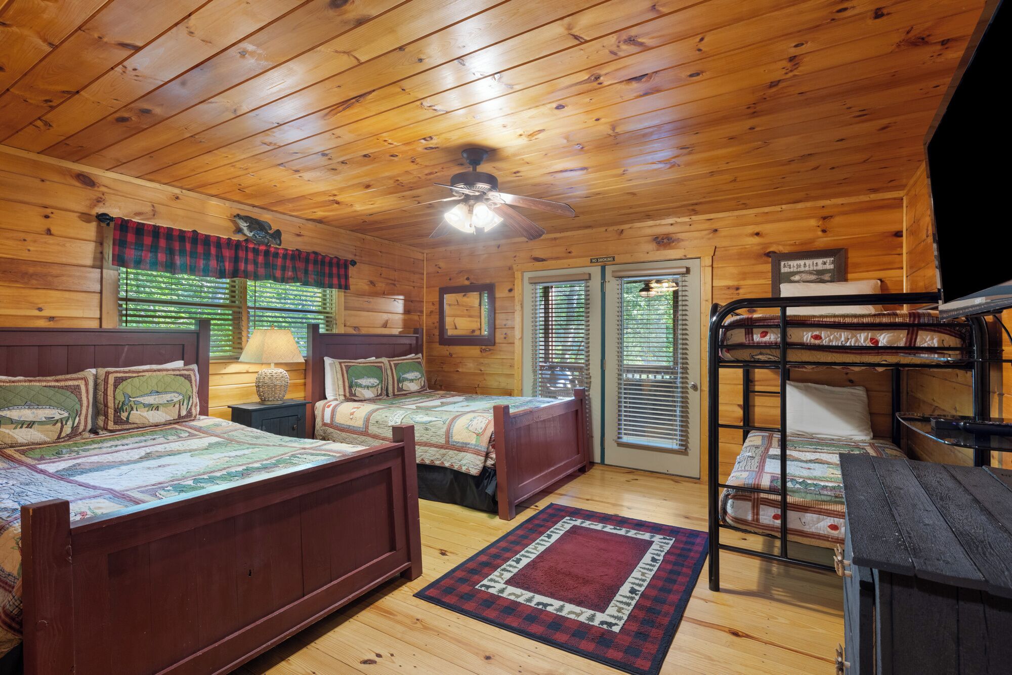 A cabin-style room with two wooden beds, a bunk bed, a ceiling fan, and a window with blinds.