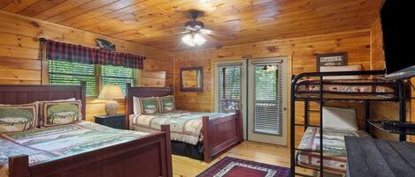 A cabin-style room with two wooden beds, a bunk bed, a ceiling fan, and a window with blinds.