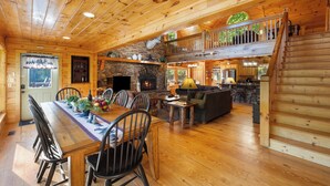 Interior espacioso de una cabaña de troncos, con comedor, sala de estar, cocina y una chimenea de piedra.