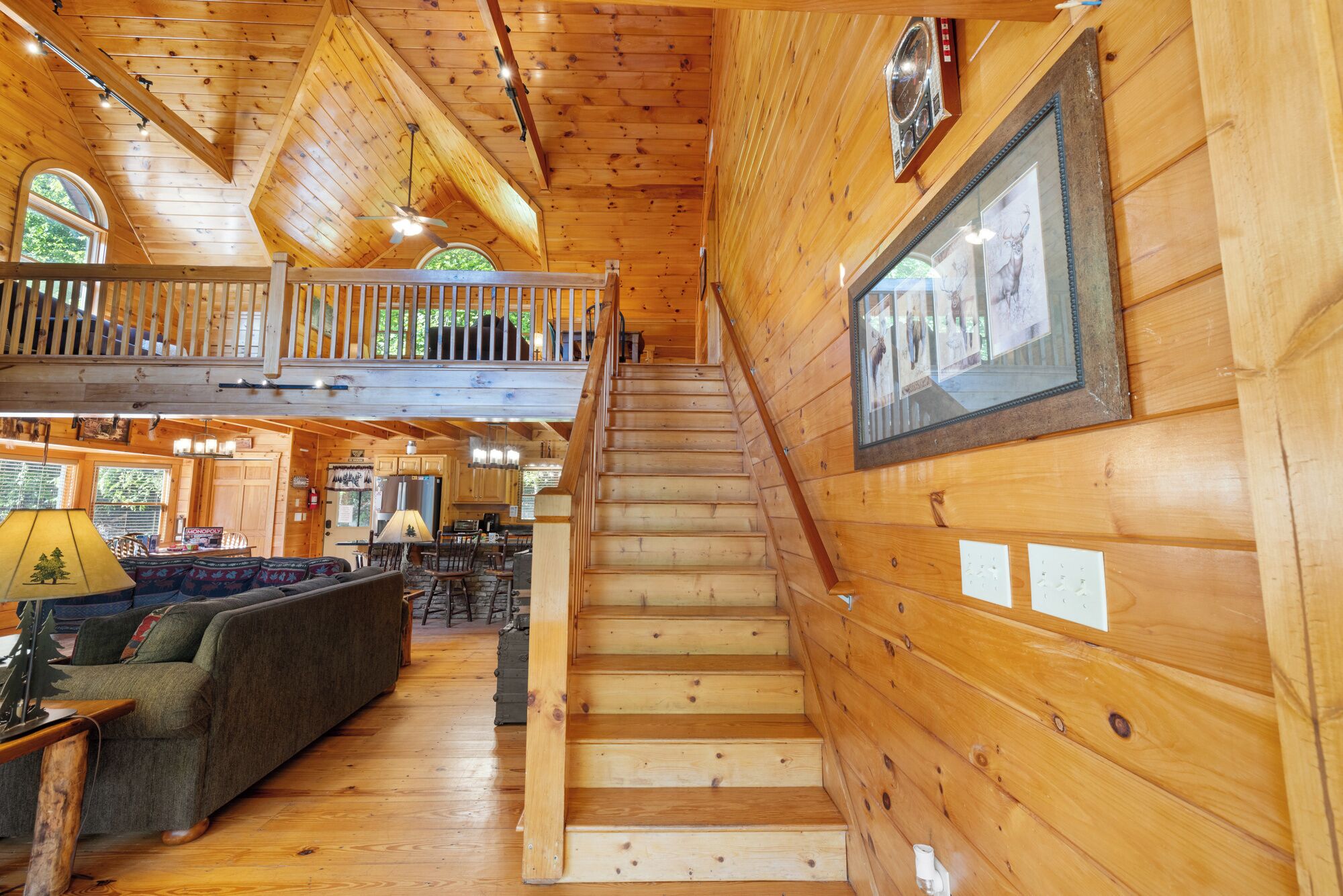 A wooden cabin interior with a staircase leading to a loft bedroom, a dining area, and a living room with a sofa and lamp.