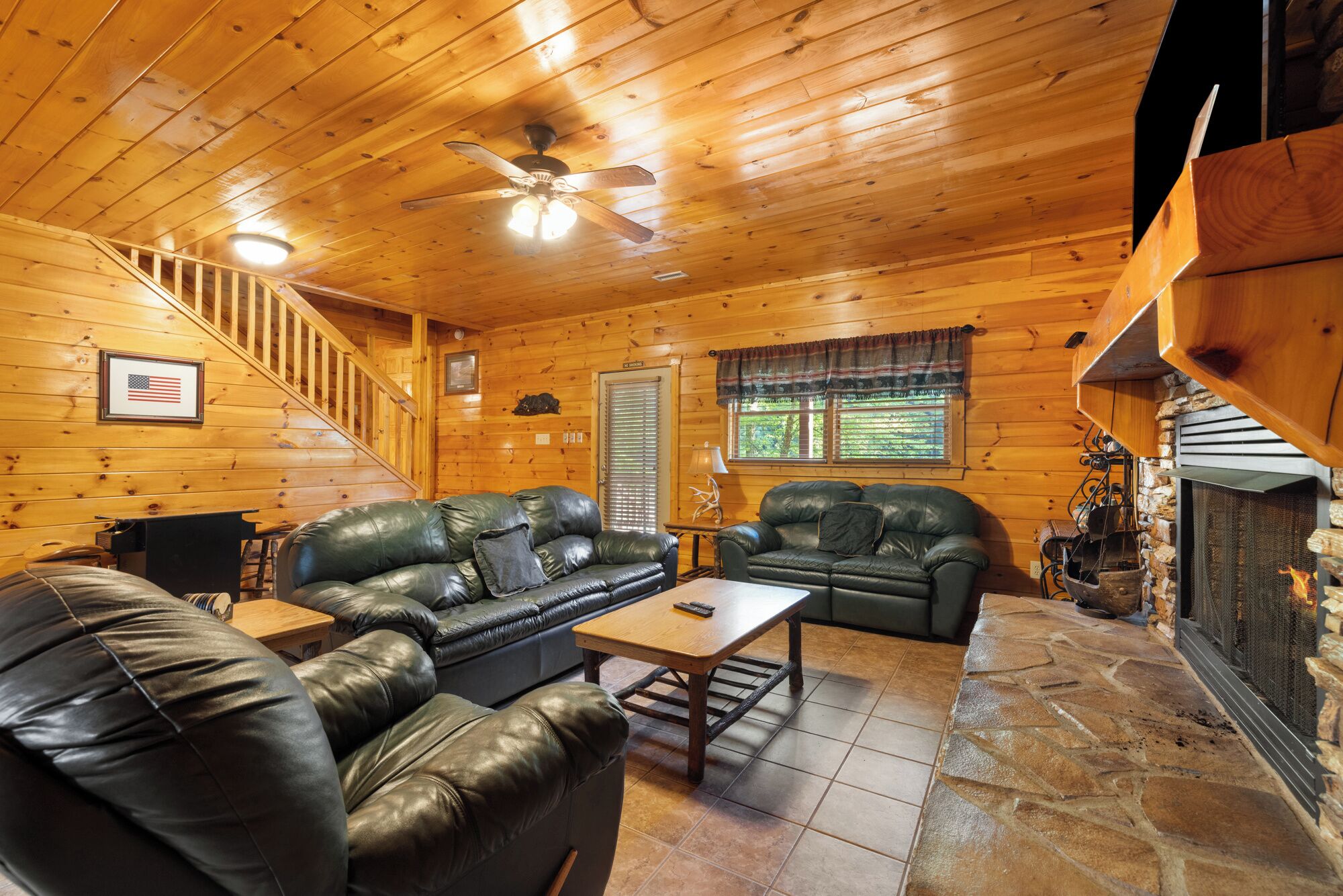 A cozy cabin interior with wooden walls, a stone fireplace, leather sofas, and a staircase.