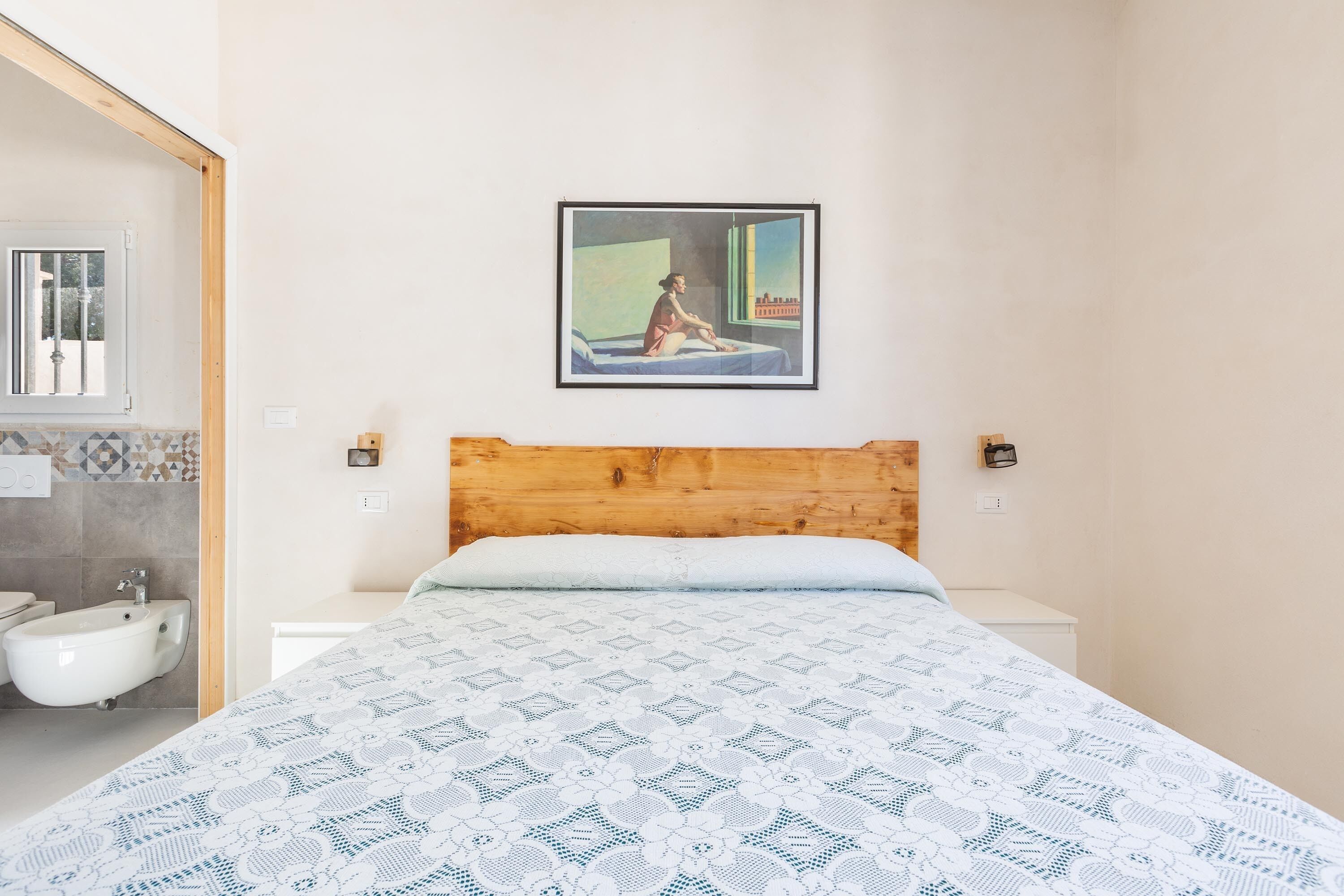 Una camera da letto con una testiera in legno, un letto con copriletto decorato, un quadro incorniciato sopra il letto e un bagno con bidet e parete rivestita di piastrelle.
