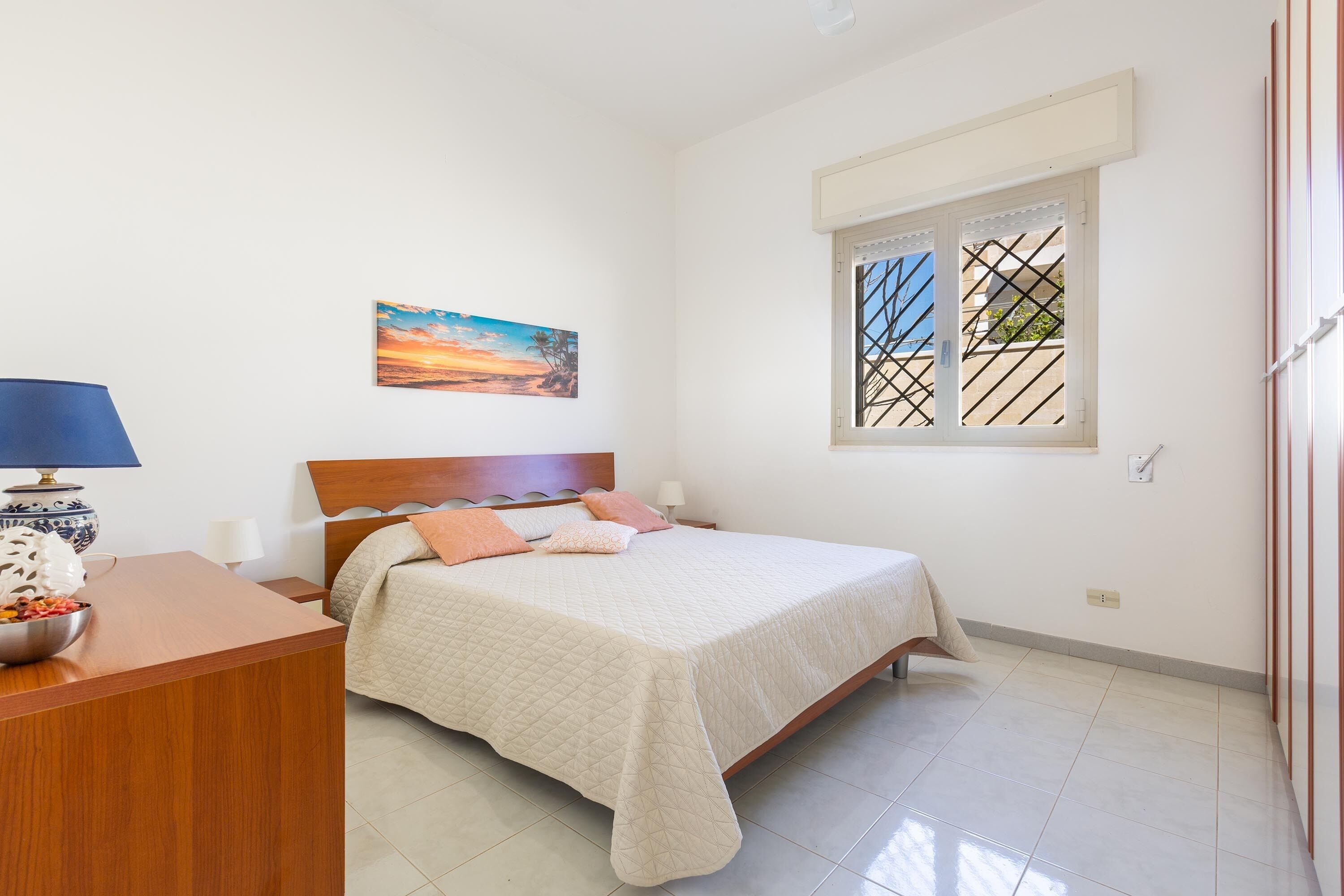 A bedroom with a bed, a nightstand, a lamp, a painting, and a window with a view.