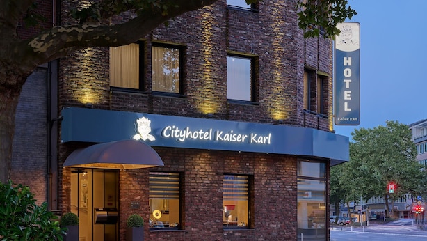 Exterior del Cityhotel Kaiser Karl, con fachada de ladrillo, ventanas amplias y un llamativo letrero iluminado.