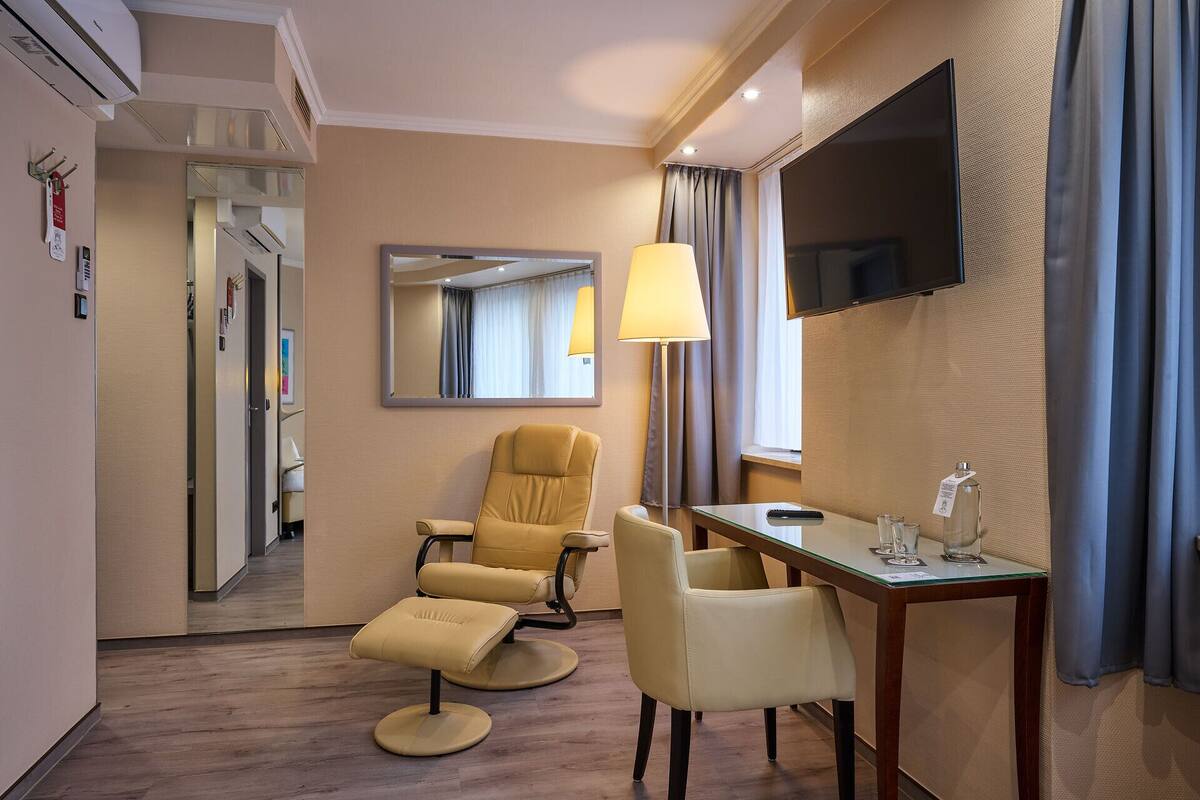 Une chambre d’hôtel équipée d’un canapé, d’un fauteuil, d’un pouf, d’un bureau, d’une télévision et d’une lampe.