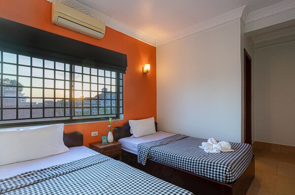 Une chambre d’hôtel avec deux lits, une grande fenêtre donnant sur un beau paysage, un climatiseur et une table de chevet agrémentée d’un vase et d’une horloge.