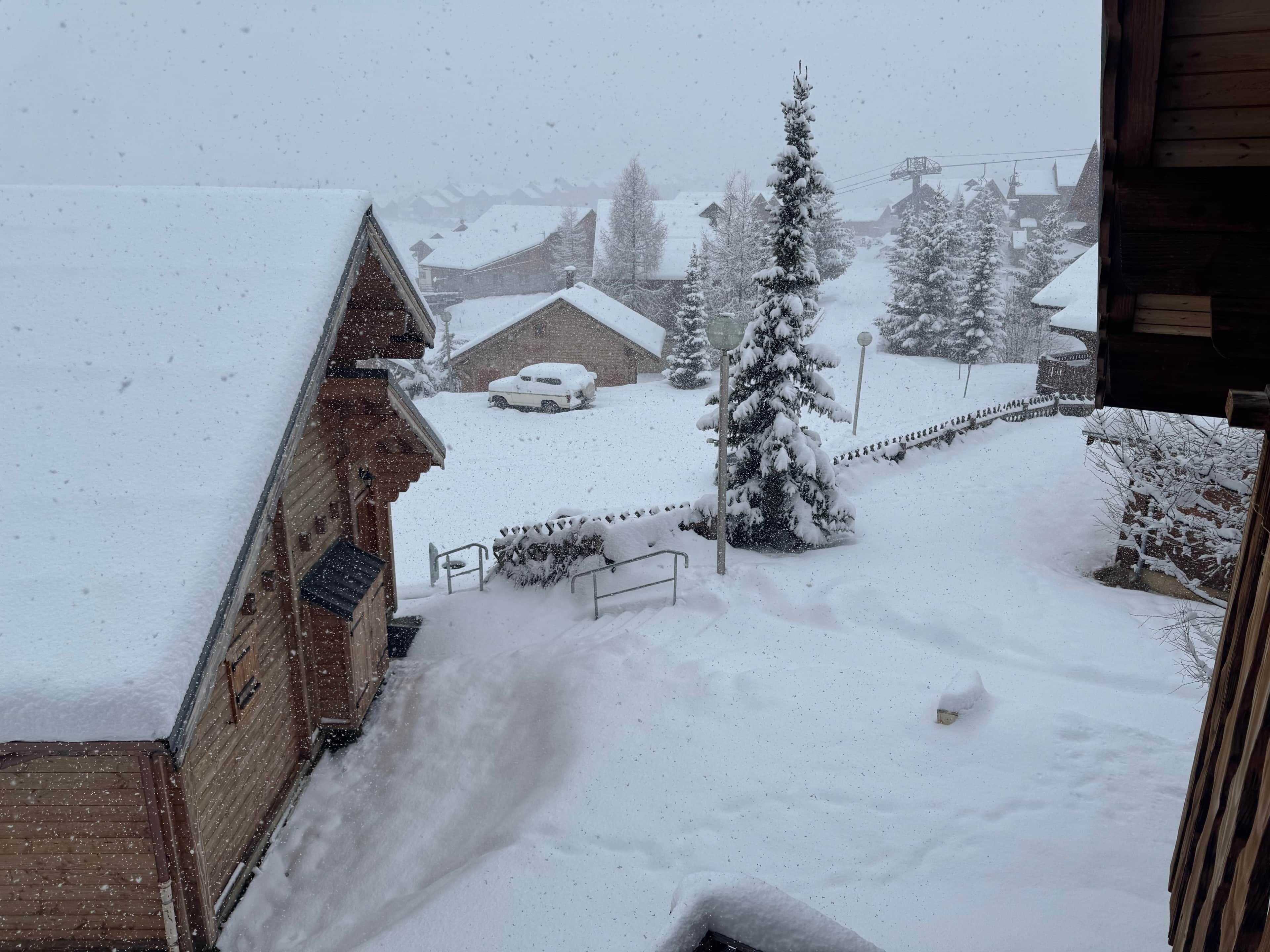 Een besneeerd landschap met een houten gebouw, een auto onder de sneeuw en een met sneeuw bedekte boom.