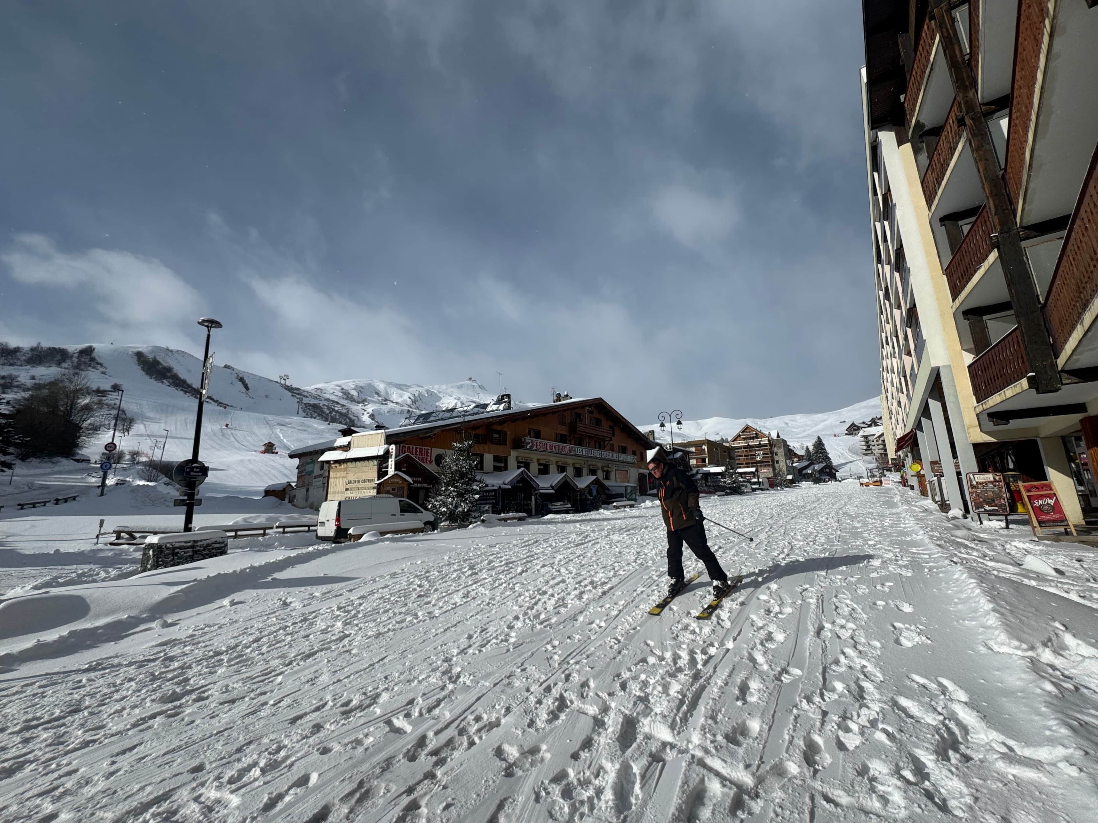 A ski resort with a person skiing on a snow-covered path, buildings with signs, and a mountain in the background.