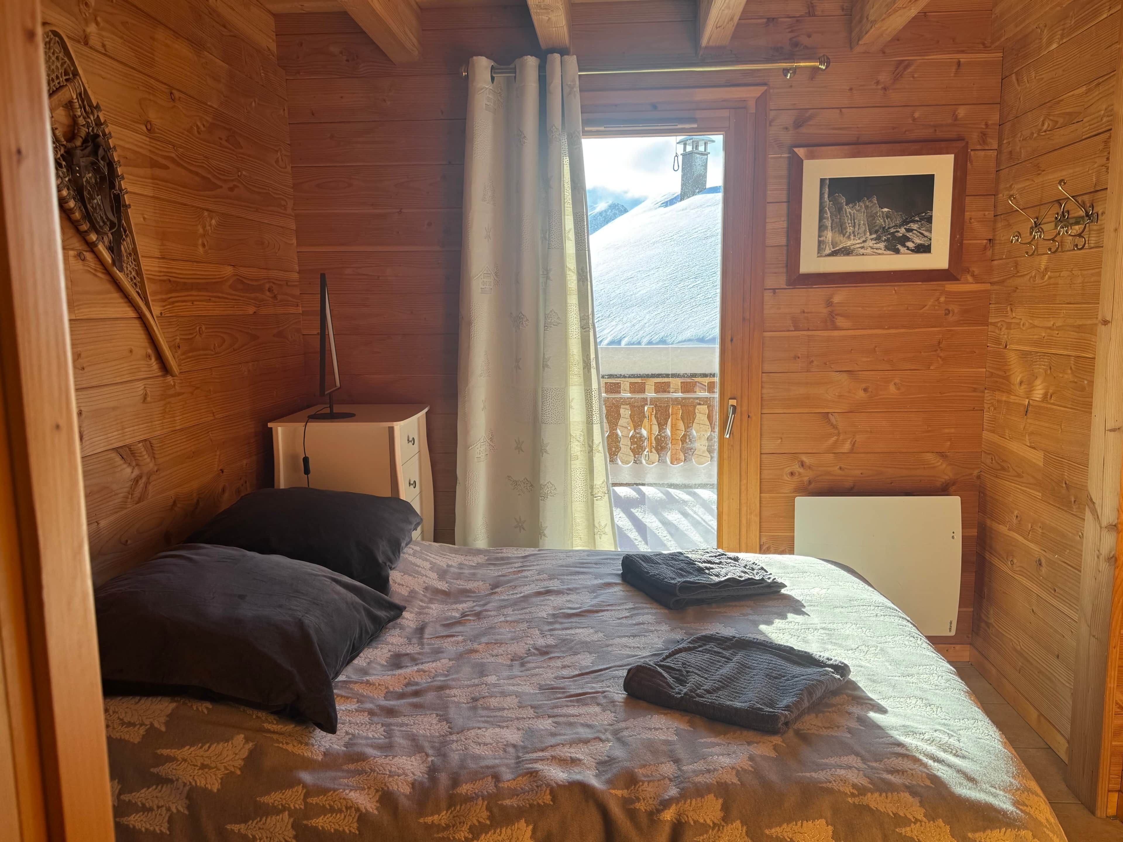 A wooden cabin room with a bed, a desk, a chair, a radiator, and a window with a view of a snowy mountain.
