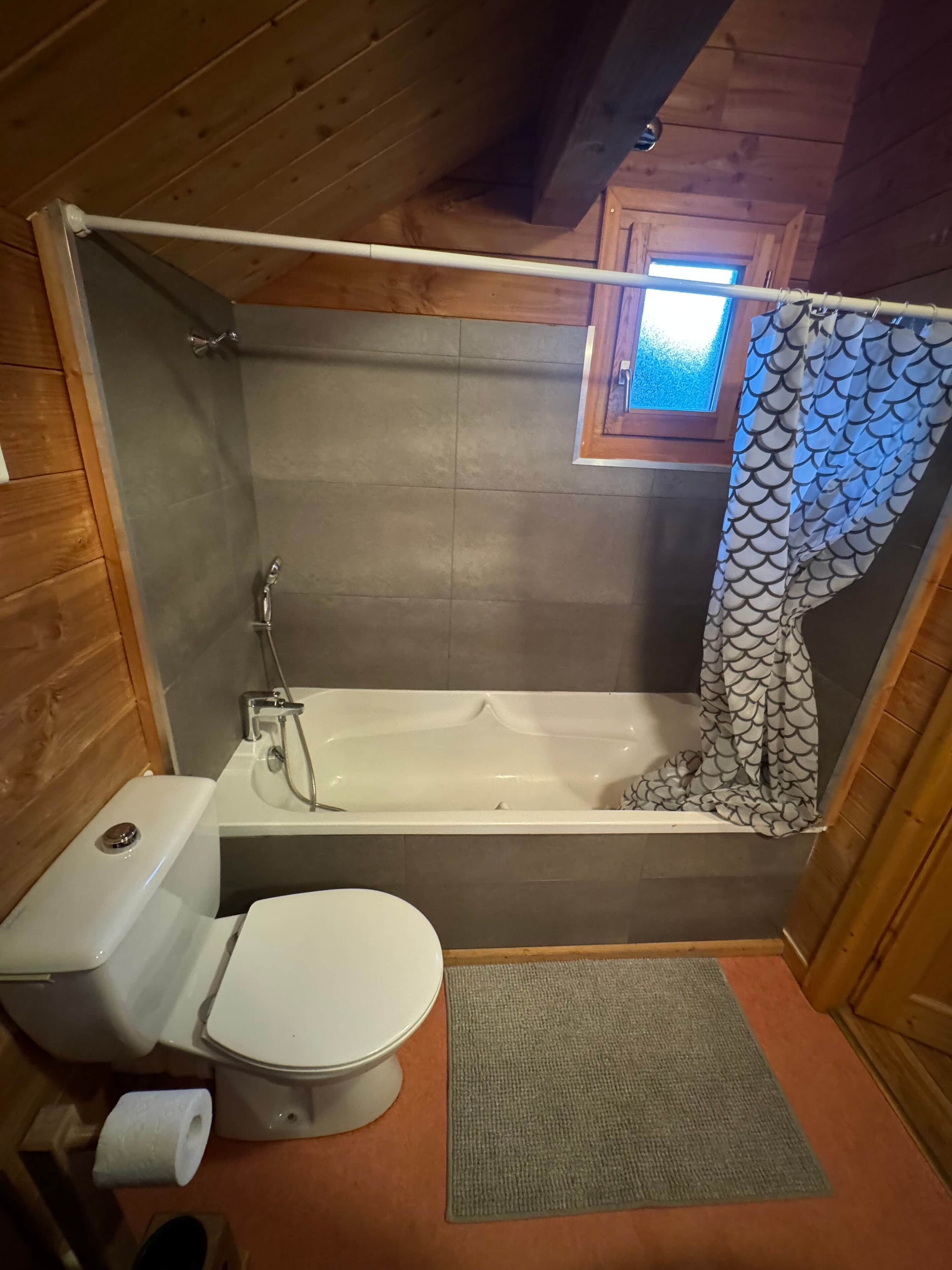 A small, wooden cabin bathroom with a bathtub, toilet, and a window above the shower.