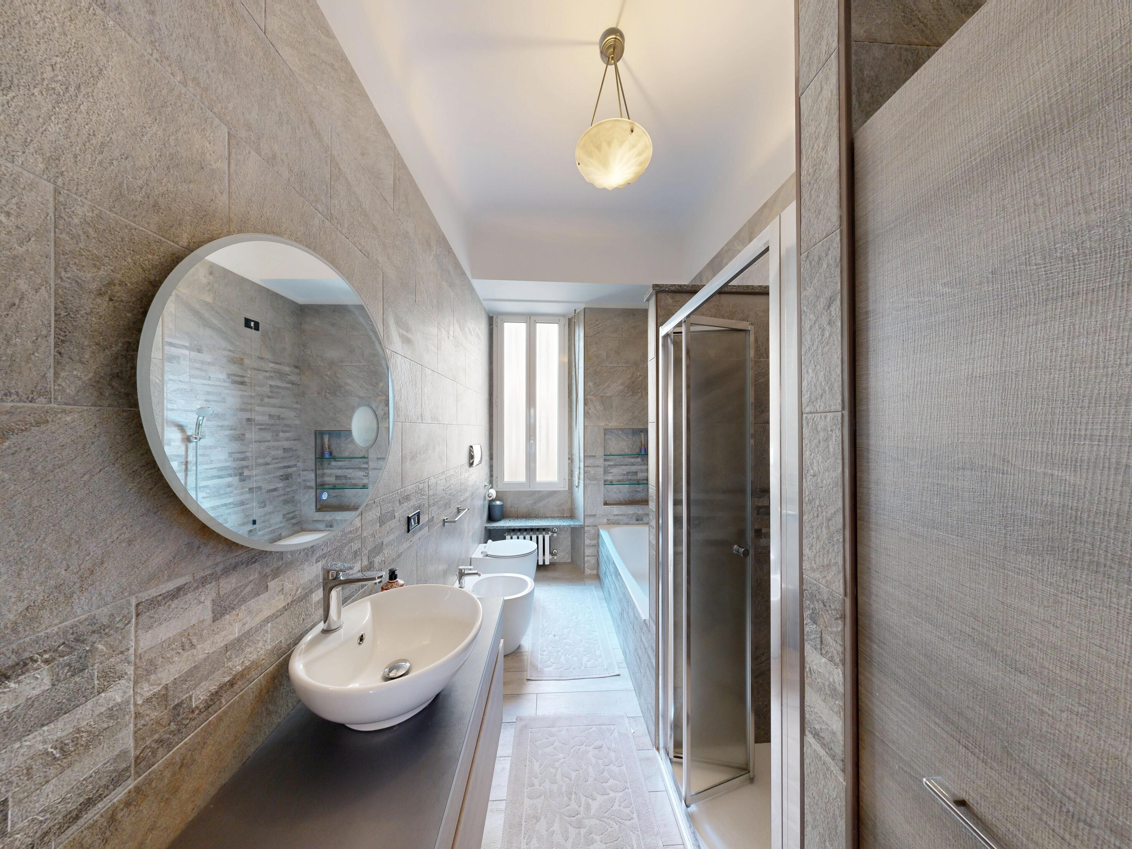 Een moderne badkamer met een grote ronde spiegel, een witte wastafel en een douche met glazen wanden.