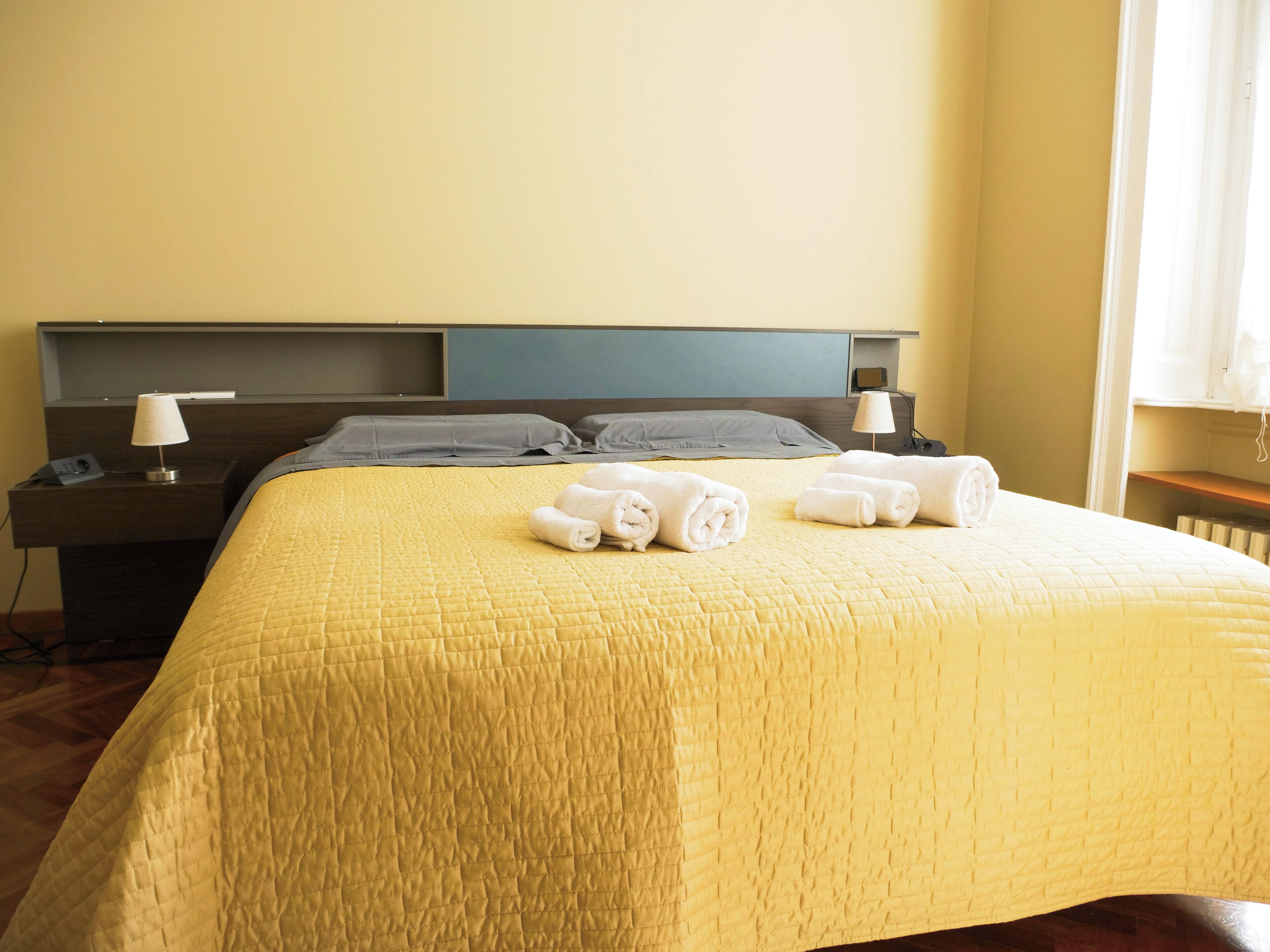 A neatly made bed with a yellow quilt, two bedside tables with lamps, and a window with curtains.