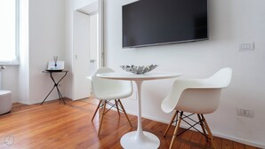 A modern living room with a round white table, two white chairs, a flat-screen TV mounted on the wall, and a small side table with a decorative bowl. - Italianway Easy - Vincenzo Monti 32 (Milan)