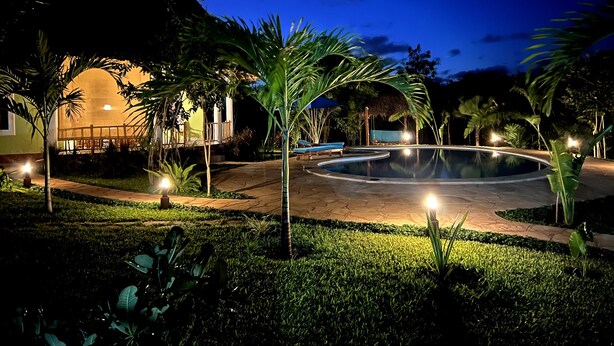 Vista nocturna de un resort con piscina, palmeras y un edificio con techo de paja.